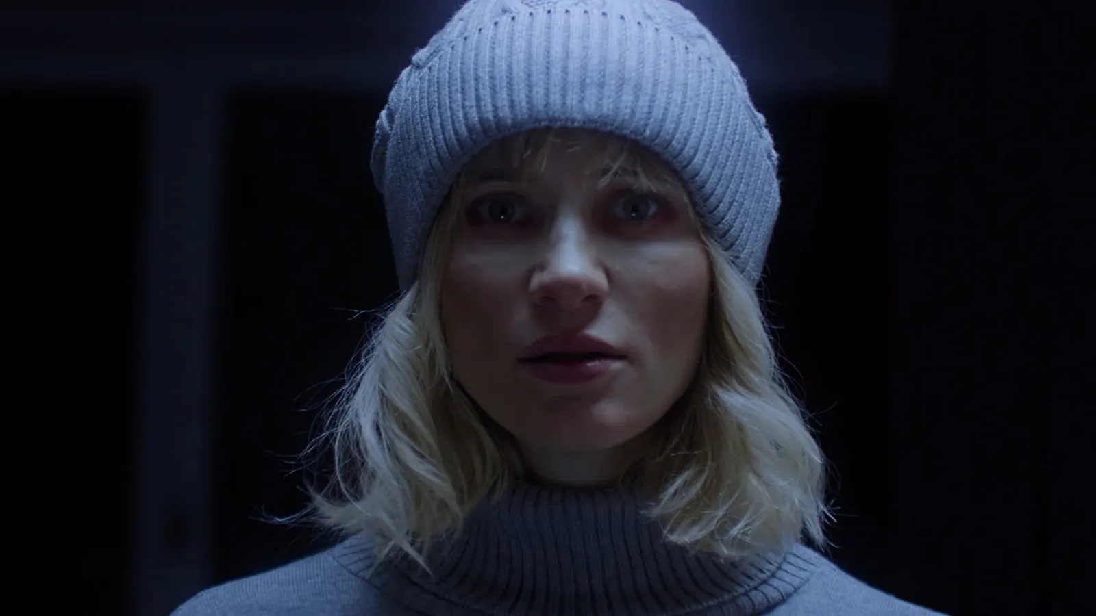 Live-action still: a woman in a grey knit hat in a dark room
